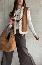 Chaleco de gamuza color camel con interior de corderito sintético, diseño sin mangas, borde frontal con ribete de corderito y detalle de tachas metálicas decorativas.