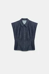 Camisa sin mangas de denim azul oscuro, con cuello solapa, hombreras marcadas, bolsillos delanteros de plastrón, bajo con peplum y cierre frontal con botones metálicos.