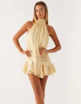 Vestido corto color beige con cuello halter alto, cuerpo holgado y falda con detalle de frunces en la cintura y ruedo con volados.