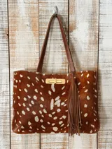 Cartera tipo tote de cuero con pelo, con estampado animal print tipo ciervo en tonos marrón y blanco. Cuenta con doble asa de hombro en cuero liso marrón, cierre frontal metálico dorado y detalle de borla decorativa lateral.