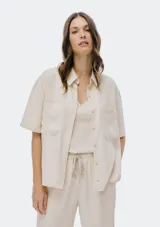 Camisa de manga corta color beige, con cuello camisero, cierre frontal de botones y dos bolsillos delanteros con solapa.