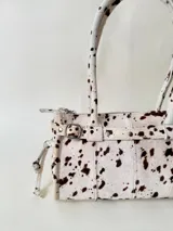 Cartera de cuero de tamaño medio con estructura rígida. Presenta una tira decorativa con tachas y hebillas metálicas en los extremos, además de lazos laterales. Cuenta con doble asa para llevar al hombro y cierre superior.