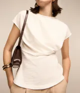 Blusa de manga corta color blanco hueso, con cuello redondo y un detalle de cruce o drapeado asimétrico en la parte delantera a la altura de la cintura. La tela presenta una textura sutil.