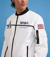 Campera tipo bomber blanca con detalles en negro, inspirada en uniformes espaciales de la NASA. Presenta parches con el logo de la NASA, la bandera de EE. UU. y texto en la manga que dice "Kennedy Space Center".