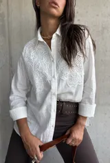 Camisa blanca de manga larga con cuello clásico y cierre frontal de botones. Presenta un diseño con paneles de encaje bordado en la parte superior delantera, confeccionada en tejido con textura.