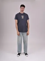 Remera gris oscuro con cuello redondo, mangas cortas con rayas blancas y amarillas en los hombros, y estampado frontal con la palabra "Brooklyn" y el número 77 en amarillo.