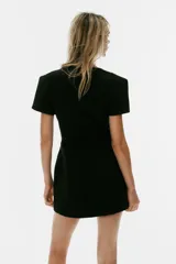 Vestido mini negro con cuello redondo, manga corta con pliegues y costuras marcadas. Cuenta con cierre posterior con cremallera oculta.
