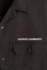 Camisa cargo de manga corta color marrón oscuro, con cuello abierto, botones frontales, bolsillos con solapa en el pecho y el texto 'SERVICE GARMENTS' bordado sobre el bolsillo izquierdo.