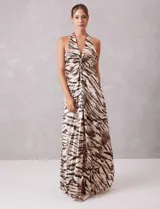 Vestido largo de tejido satinado plisado con estampado animal print tipo cebra en tonos tierra y ocre. Presenta escote halter profundo en V con drapeado central y broche de pedrería, espalda descubierta y falda fluida con apertura frontal.