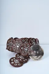 Cinto de cuero marrón compuesto por piezas circulares unidas, decoradas con apliques metálicos plateados con diseño floral y remaches.