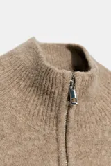 Jersey de punto de color beige con cuello subido y cierre frontal con cremallera metálica. Presenta mangas largas con puños acanalados y diseño de corte holgado.