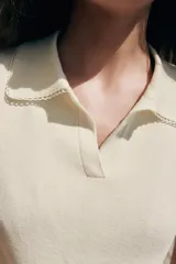 Top de punto color crudo, con cuello polo y escote en V. Cuenta con mangas cortas y detalle de puntilla con forma de onda en el cuello y los puños.