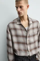 Camisa relaxed fit de manga larga con cuello solapa, confeccionada en tejido con mezcla de viscosa. Presenta un bolsillo plastrón en el pecho, bajo con aberturas laterales y cierre frontal de botonadura. El diseño es a cuadros en tonos beige y marrón.