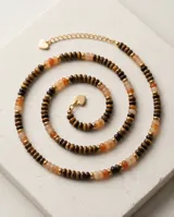 Collar de acero con terminaciones en baño de oro y cuentas de madera y piedras naturales en tonos marrones y naranjas, con cierre de mosquetón y cadena de alargue con dije de corazón.