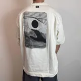 Remera de corte oversize confeccionada en algodón pesado, color blanco. Presenta un parche rectangular en la espalda con una estampa artística en blanco y negro que muestra una mano alcanzando un círculo oscuro sobre un fondo de líneas horizontales.