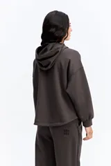 Hoodie de algodón color marrón con capucha, cierre frontal parcial y detalle de costuras horizontales en contraste negro. Presenta diseño holgado, mangas largas con puños elásticos y cordones ajustables en la capucha.