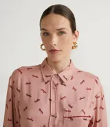 Camisa de viscosa color rosa con estampado de caballos en marrón. Tiene cuello camisero, cierre con botones, mangas largas, un bolsillo frontal y detalle de vivo contrastante en el bolsillo.