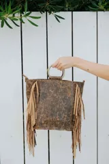 Cartera de cuero con acabado envejecido y flecos laterales en color beige. Presenta asas circulares metálicas y un diseño estructurado.