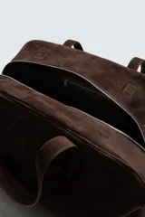 Bolso de viaje de gran tamaño confeccionado en piel con acabado de serraje. Cuenta con un compartimento principal con cierre de cremallera e imanes, interior con bolsillo con cierre y bolsillo plano, y dos asas de mano de piel.
