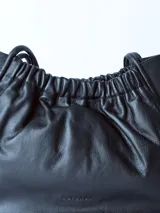 Bolso tipo tote bag de cuero negro, con diseño fruncido en la parte superior y dos asas delgadas de hombro. Presenta un acabado de cuero suave y arrugado.