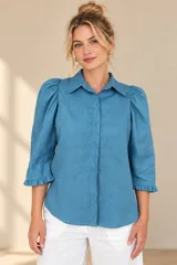 Camisa de lino color blanca, con cuello clásico, mangas tres cuartos abullonadas en los hombros y terminación con volados en los puños, cierre frontal con botones ocultos.