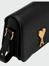 Clutch de cuero negro con solapa frontal y cierre magnético. Presenta el icónico logo metálico dorado en forma de corazón con la letra A en el centro. Incluye una correa fina ajustable y extraíble para llevar al hombro o cruzada.