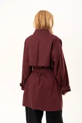 Trench de corte holgado color bordó, con cuello camisero, cierre frontal con botones y cinturón ajustable con hebilla. Presenta mangas largas con detalles de correas en los puños y bolsillos laterales.