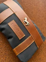 Sobre tipo cartuchera de lona naranja con detalles y apliques en cuero marrón. Presenta un aplique metálico en forma de caballo y un parche con el logo de la marca grabado en el frente.