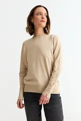 Sweater de punto color beige, con cuello redondo, manga larga y puños con abertura lateral decorados con pequeños botones metálicos.