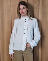 Camisa de algodón color blanco, con cuello alto y volados laterales que recorren la botonadura frontal. Presenta mangas largas con puños elásticos terminados en volados.