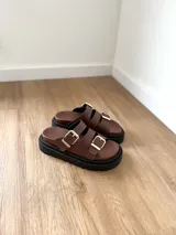 Sandalias marrones de cuero con doble tira y hebillas doradas, sobre base chunky negra.