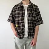 Camisa de manga corta de corte boxy fit, con estampa a cuadros en tonos marrones, azules y blancos. Cuenta con cierre frontal de botones y un bolsillo ojal en el pecho.