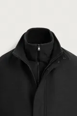 Abrigo negro de corte recto con cuello subido y doble cuello desmontable mediante cremallera interior. Cuenta con manga larga, bolsillos de vivo en la cadera, bolsillo interior, abertura central en la espalda y cierre frontal de cremallera oculta por solapa con botones a presión.