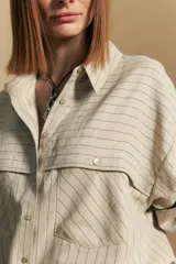 Camisa de manga larga color blanco hueso con finas rayas verticales en color oscuro. Presenta cuello camisero, botones frontales y dos bolsillos superpuestos en el pecho.