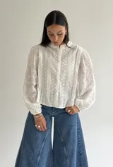 Blusa de brodery color blanco con cuello redondo, mangas largas abullonadas con puños elásticos y cierre frontal con botones forrados. Presenta detalles de puntillas verticales y calados en toda la prenda.