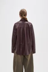 Sobrecamisa de cuero sintético en tono bordó oscuro, con cuello clásico, cierre frontal oculto y un bolsillo aplicado en el pecho. Presenta un corte holgado con mangas largas y puños rectos.