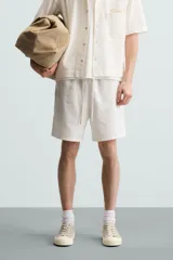 Bermuda de algodón color blanco, de corte relajado (relaxed fit), con cintura elástica ajustable mediante cordón. Posee bolsillos laterales y un bolsillo trasero de plastrón. Presenta un bordado en contraste en la pernera.