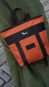 Mochila pequeña color borgoña con detalles en cuero marrón y aplique metálico de un caballo. Tiene manija superior y correas traseras regulables.