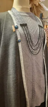 Capa larga gris con vivo blanco en los bordes y botones decorativos. Se cierra con broche en el cuello.