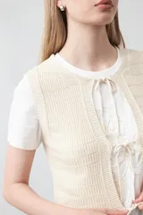 Chaleco tejido color verde musgo, con trama texturada, cuello a la base y cierre frontal con lazos. Presenta un ruedo con volado.