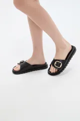 Sandalia plana de eco cuero negro, con tira ancha en el empeine decorada con una hebilla metálica dorada cuadrada y suela dentada.