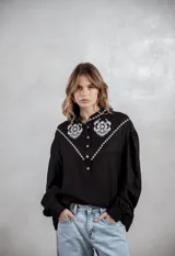 Camisa negra de corte holgado con mangas largas abullonadas y puños abotonados. Presenta un diseño con cuello mao, cierre frontal parcial con botones y detalles de bordado floral en color blanco en la zona del pecho y hombros.