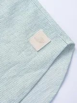 Camisa de manga corta confeccionada en tejido ligero de algodón con diseño de rayas verticales finas. Presenta cuello clásico, cierre frontal con botones de madera, un bolsillo aplicado en el pecho y un pequeño parche decorativo en el ruedo.