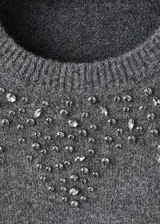 Jersey de punto grueso color gris, con cuello redondo, manga larga y diseño recto. Presenta un detalle decorativo de cristales en la zona de los hombros y el pecho.