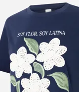 Remera de algodón color blanco, con cuello redondo y manga corta. Presenta un estampado con la frase 'Soy flor. Soy latina' y flores bordadas en relieve con textura tejida.