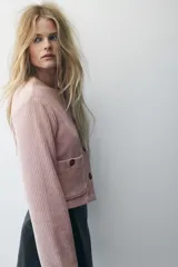 Chaqueta corta de velour acanalado color rosado, con cuello redondo, cierre frontal de botones marrones y dos bolsillos de parche con botón en la parte delantera. Presenta hombros caídos y mangas largas.