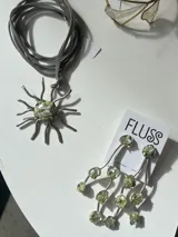 Collar artesanal de la marca Fluss con dije en forma de sol hecho de bronce y alpaca, con incrustaciones de piedra en tono verde y centro de resina con fragmentos. Viene con una tira de gamuza gris.