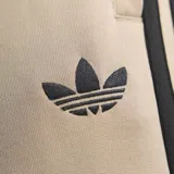 Pantalón de jogging Adidas de corte regular en color beige, con las tres tiras características en negro a lo largo de los laterales y el logo del trifolio bordado en la parte frontal.