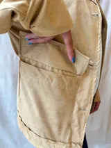 Chaqueta color beige con cuello redondo, cierre frontal con botones a presión, bolsillos de parche y mangas largas.