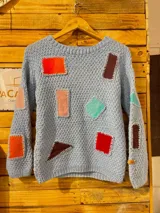 Buzo celeste de punto tejido a mano, con mangas largas y cuello redondo, decorado con apliques geométricos de cuero en diversos colores unidos con puntadas visibles.